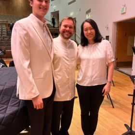 Clancy in a white suit jacket, Marcelo, and Heidi smiling together on an auditorium stage at a Western Washington University music event.