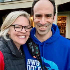 Erika and Ira smiling together at a community event, wearing a Northwest Branch NWB Dolphins blue hoodie and a black puffer vest.