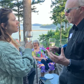 Don and Caroline holding wine glasses and talking on a scenic deck overlooking Chuckanut Bay, with Phyllis Self seated in the background.