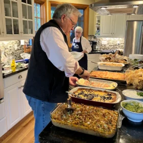 Charlie serving fresh salmon from a kitchen buffet next to Mary Pat during a Bellingham Festival of Music dinner party.