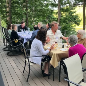 Guests enjoying an outdoor dinner on a wooden deck surrounded by evergreen trees at a Bellingham Festival of Music sponsor party.