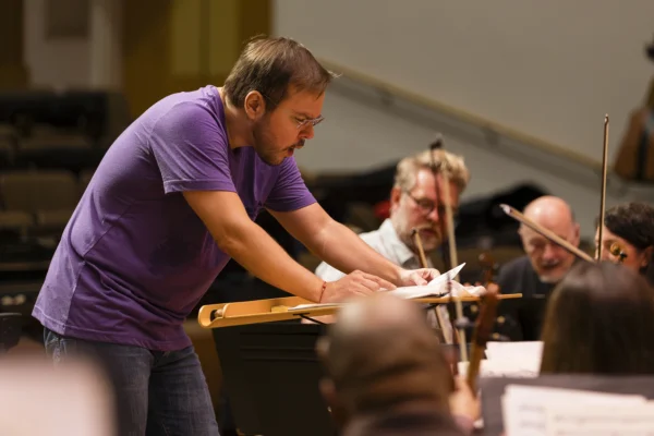 Conductor Marcelo Lehninger leaning over a music stand score while directing orchestra musicians in a purple rehearsal shirt.