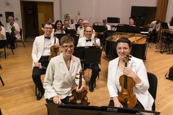 Orchestra string players seated in formal white jackets posing with violins on stage before a concert performance.