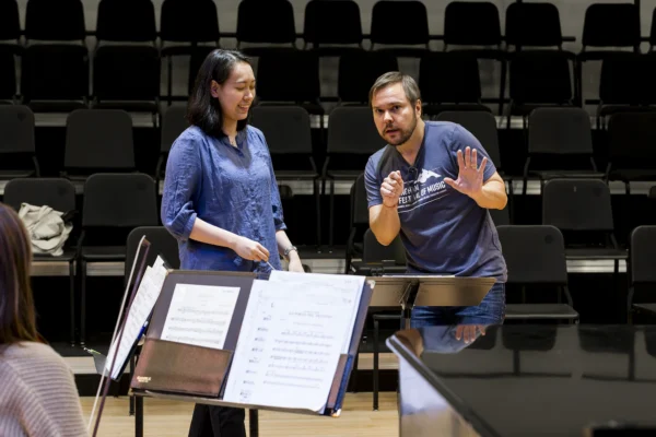 Conductor in a festival t-shirt coaching a female student conductor in front of a music stand and piano during a conducting masterclass.