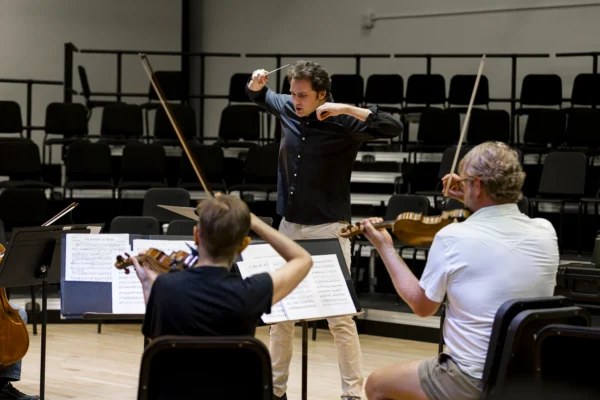 Male student conductor leading an ensemble of string players with intense focus during a conducting workshop.