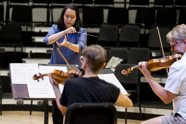 Female conductor holding a baton and using expressive hand signals to lead string musicians during a music workshop or rehearsal.
