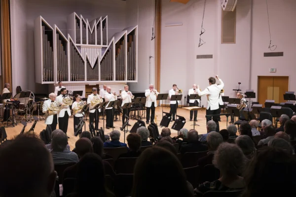 Conductor leading a full brass section performing standing on stage beneath pipe organ pipes during a concert.