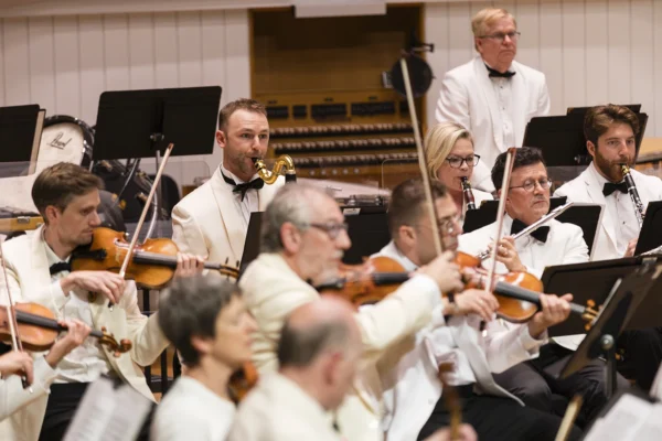Orchestra musicians in formal white jackets playing violins, woodwinds, and percussion during a live summer festival concert.