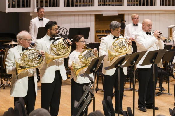 Brass section featured on stage, including French horn and trumpet players performing in formal white dinner jackets during a symphony concert.