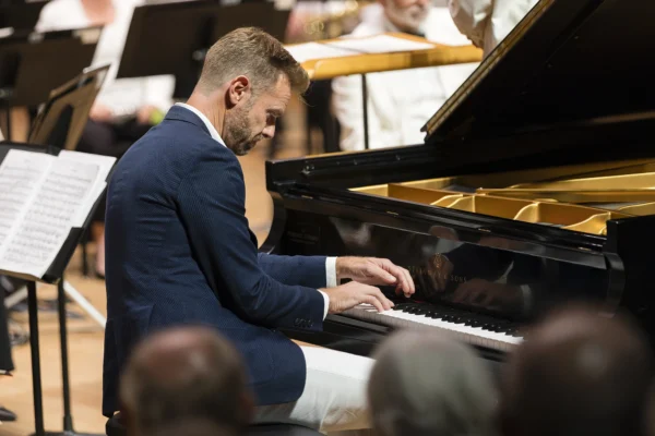 Male piano soloist Andrew von Oeyen playing a grand Steinway piano on stage backed by an orchestra during a concerto performance.