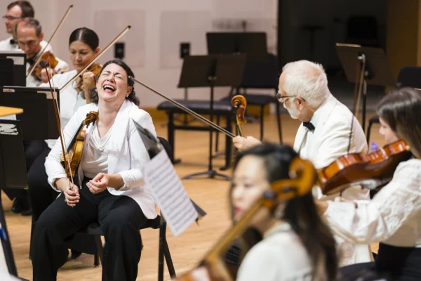 Female violinist in a white jacket laughing joyfully during a playful lighthearted moment on stage with the orchestra.