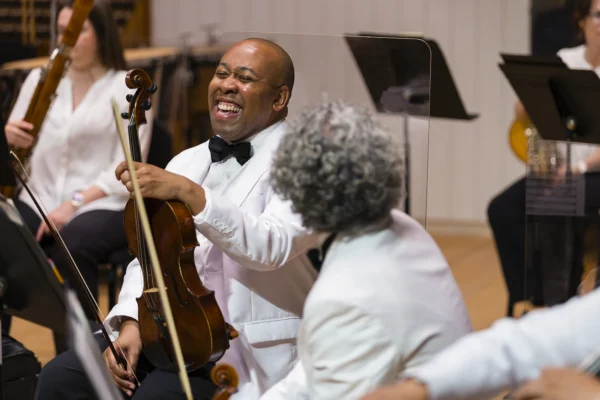 Smiling male viola player in a white dinner jacket laughing while seated in the string section during a concert break.