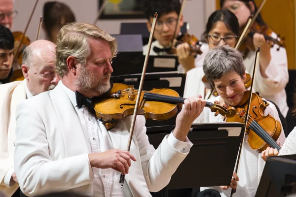 Male violinist in a white dinner jacket playing an intricate section while seated among orchestra string musicians.