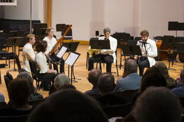 Chamber ensemble featuring woodwind and French horn musicians performing on stage for an audience.