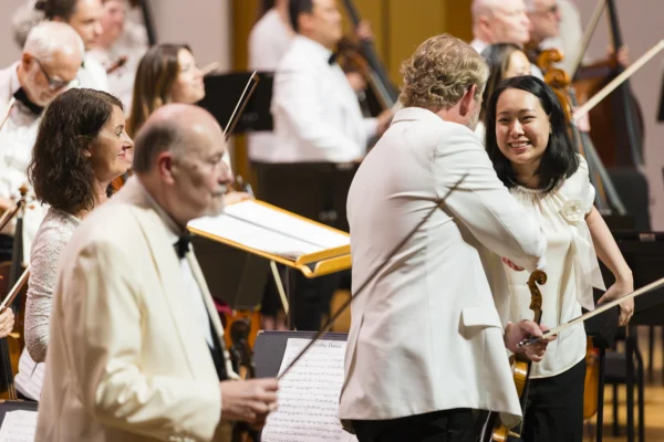 Violinist in a white jacket shaking hands with a smiling female musician on stage during applause.
