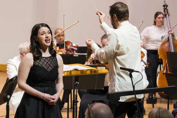 Soprano vocal soloist Maria Valdes in a black evening gown singing on stage alongside an orchestra conductor.