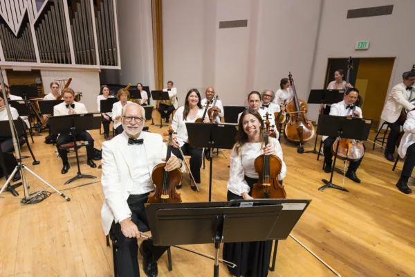 Seated violin and string section musicians smiling on stage in formal white jackets prior to a performance.