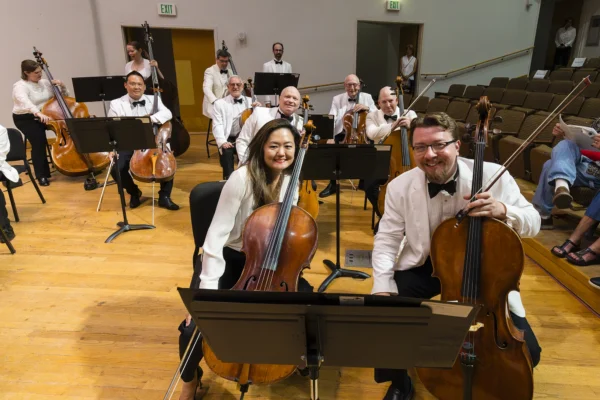 Two cellists seated in the front row of an orchestra smiling toward the camera with their instruments.