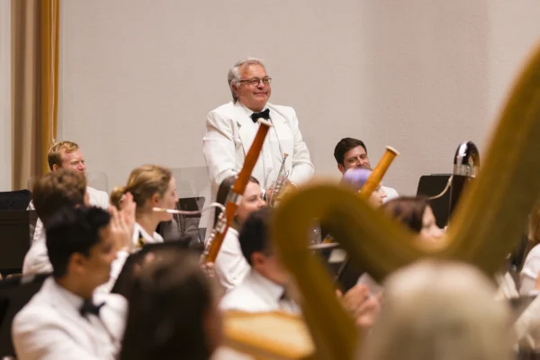 Male trumpet player in a white dinner jacket standing to take a bow while fellow musicians applaud.