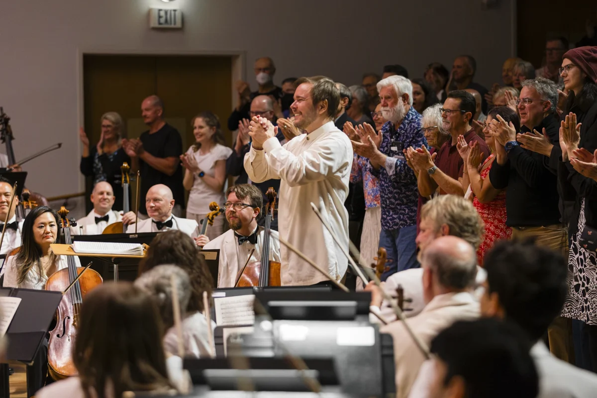 Conductor Marcelo Lehninger applauding toward a standing audience during a standing ovation at the end of an orchestral concert.