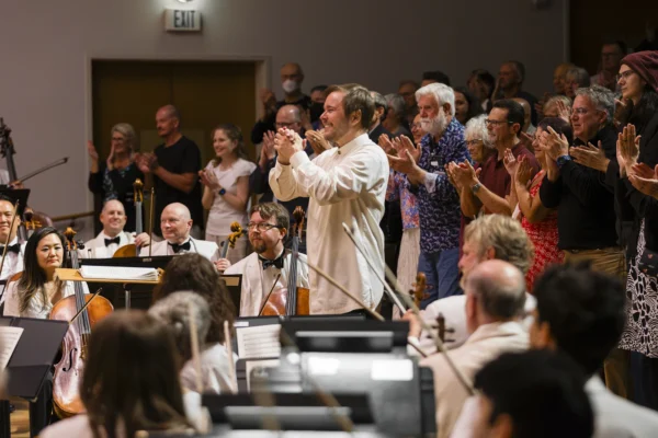 Conductor Marcelo Lehninger applauding toward a standing audience during a standing ovation at the end of an orchestral concert.