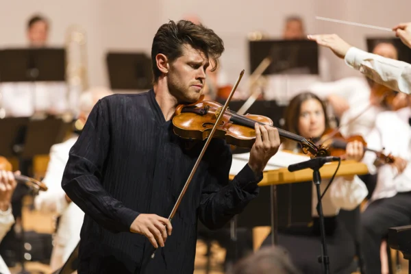 Guest violin soloist Blake Pouliot in a dark shirt performing intently with a microphone set up near his instrument.