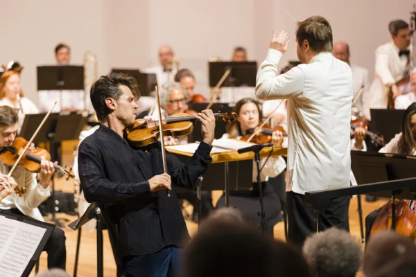 Violin soloist Blake Pouliot playing on stage alongside a standing conductor directing the orchestra.