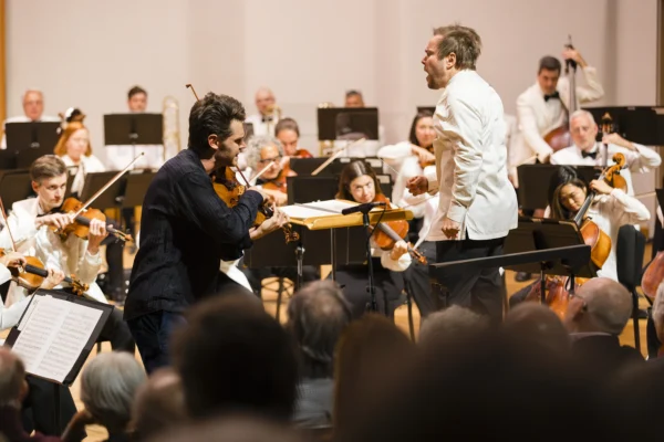 Conductor gesturing dramatically toward male violin soloist Blake Pouliot during an intense passage of a concerto performance.